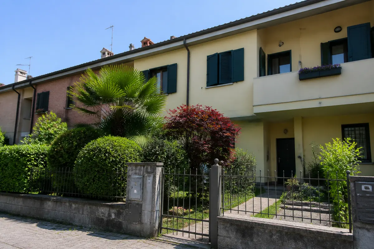 Villetta a schiera con giardino verde, cancello e facciate colorate illuminate dal sole.